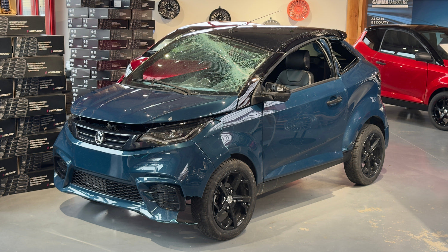 Verunfalltes Leichtkraftfahrzeug (Aixam) mit eingedrücktem Dach und gesplitterter Frontscheibe in einer Ausstellungshalle – zeigt die Stabilität der Fahrgastzelle nach einem Überschlag.